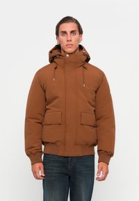 Brązowa pikowana kurtka z kapturem, z dwiema przednimi kieszeniami, sznurkiem do regulacji i ściągaczem na dole. Gładka tekstura i jednolity kolor.