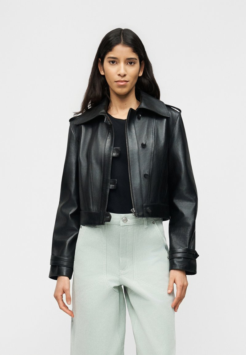 Γυναίκα με μακριά σκούρα μαλλιά, φοράει κοντό μαύρο δερμάτινο μπουφάν πάνω από μαύρο τοπ και ανοιχτό γκρι ψηλόμεσα παντελόνια φαρδιάς γραμμής, όρθια.