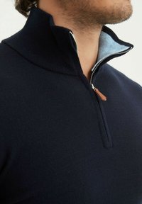 Pull zip en navy avec col montant, tissu doux à texture côtelée et doublure intérieure contrastante en bleu clair. Détail en cuir sur la tirette.