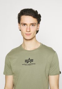 Oliven grønn t-skjorte med korte ermer laget av bomull, med en svart trykt logo på brystet. Glatt tekstur med en klassisk passform.