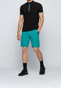 Černý top s krátkým rukávem a tyrkysovými akcenty, s půlzipem u límce, doplněný tyrkysovými šortkami, které mají kapsy a elegantní design. Černé tenisky.