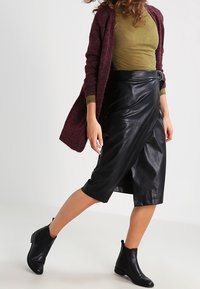 Svart läderomslutningskjol med hög midja och sidoslits, matchad med en figursydd olivgrön topp och en mörk vinröd cardigan. Svarta ankelskor.