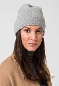 Beanie grigia lavorata a maglia con una finitura morbida e texture, caratterizzata da un orlo arrotolato. Indossata con un dolcevita scuro e un cappotto beige chiaro.