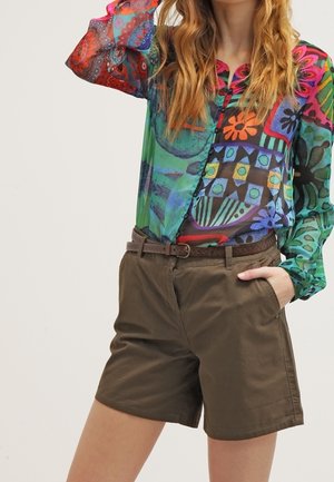 Woman wearing a colorful patterned sheer blouse tucked into olive green belted shorts with one hand in pocket.
