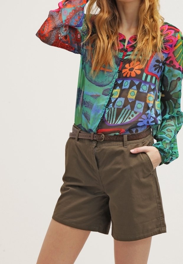 Femme portant un chemisier transparent coloré à motifs rentré dans un short vert olive ceinturé, une main dans la poche.