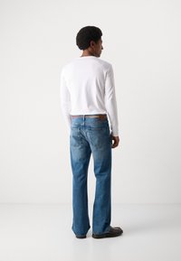 Blaue Jeans aus Denim mit geradem Bein, sichtbaren Gesäßtaschen und einem braunen Lederpatch an der Taille. Kombiniert mit einem weißen Langarmshirt.