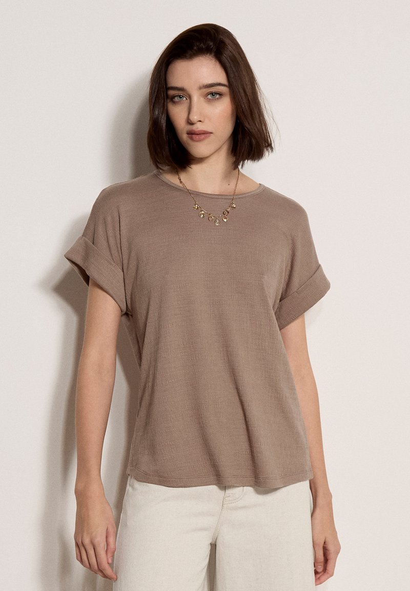 Jeune femme portant un haut taupe ample à manches courtes retroussées, un pantalon blanc et un collier pendentif en or, debout devant un fond uni.