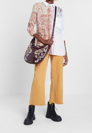 Personne portant une chemise bicolore à motifs, un pantalon court jaune, des bottes noires et portant un sac violet foncé avec une broderie florale.