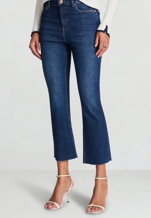 Jeans corti in denim scuro con orlo svasato, caratterizzati da una chiusura frontale con bottoni e sottili dettagli di cucitura. Indossati con tacchi bianchi con cinturini.