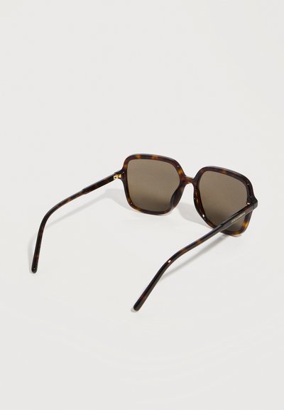 Τετράγωνα γυαλιά ηλίου με φαρδιά καφέ κορνίζα τύπου χελώνας, σκουρούς φακούς και λεπτούς βραχίονες, τοποθετημένα σε λευκή επιφάνεια.