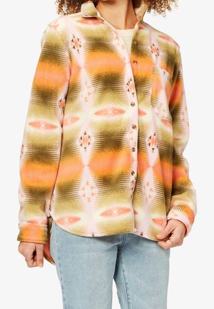 Persoon die een oranje, groene en crèmekleurige fleece-overhemd met patroon draagt, met knopen dicht, gecombineerd met lichtblauwe spijkerbroek, en het zoom van het overhemd vasthoudt.