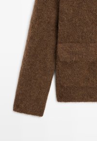 Dettagli in lana marrone pelosa sulle maniche e sulla tasca di un maglione lavorato a maglia, mostrato su uno sfondo bianco.