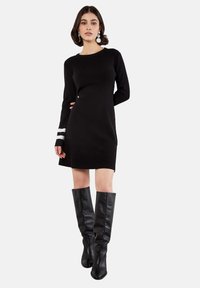 Schwarzes Strickkleid mit langen Ärmeln; verfügt über weiße horizontale Streifen an den Bündchen. Kombiniert mit hohen schwarzen Lederstiefeln.