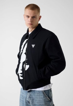 Młody mężczyzna z krótkimi włosami w czarnej kurtce zapinanej na zamek z białą grafiką twarzy i jasnoniebieskich dżinsach, ręce w kieszeniach, neutralne tło.