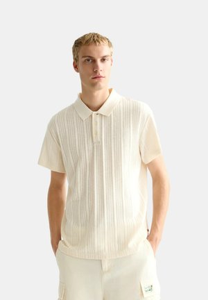 Young man with short blond hair wearing a light beige short-sleeve polo shirt and matching beige cargo shorts against white background.