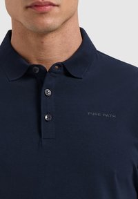 Marineblauwe katoenen polo met een klassieke kraag, drie knopen en een lichte glans. Voorzien van het "PURE PATH" logo op de borst.