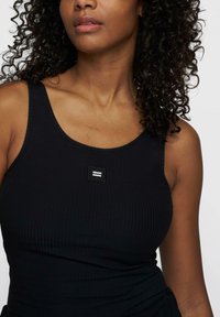 Top de tirantes negro con canalé que presenta un pequeño parche cosido con dos líneas horizontales. La tela es ajustada y se estira para mayor comodidad.
