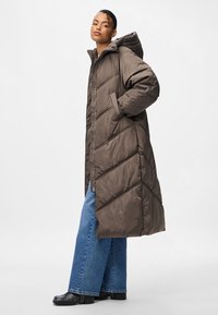 Ruda ilga pūkinė striukė su kapišonu, dekoruota įstrižinėmis siūlėmis ir šoninėmis kišenėmis. Derinama su šviesiai mėlynais plačiomis kelnėmis ir juodais batais.