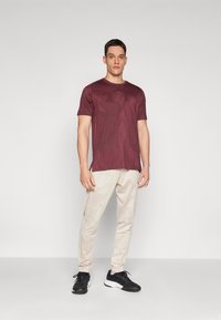 Camiseta de manga corta burdeos con un patrón geométrico; combinada con pantalones claros de color beige con estampado y zapatillas deportivas negras.