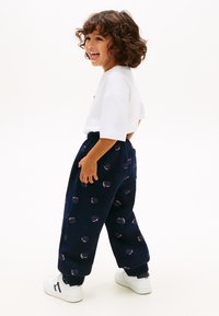 Χαμογελαστό παιδί με σγουρά μαλλιά φοράει λευκό μπλουζάκι, μπλε σκούρη φόρμα με σχέδια καλαθοσφαίρισης και λευκά αθλητικά παπούτσια, στέκεται πλάγια σε λευκό φόντο.