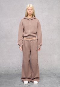 Sudadera con cremallera de forro polar marrón, con puños acanalados, combinada con pantalones jogger de pierna ancha. Ambos tienen una textura suave, presentan un corte casual y un branding mínimo.