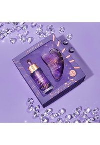 Un emballage violet avec des accents dorés contient une bouteille en verre avec un compte-gouttes et un outil en améthyste en forme de cœur. Des gemmes transparentes entourent le tout sur une surface violet clair.