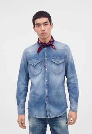 Ung mand iført en falmet blå denimskjorte med forlommer, et rødt ternet halstørklæde og slidte jeans, stående foran en ensfarvet baggrund.