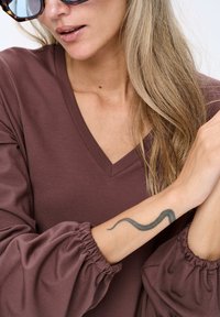 Vrouw met lang blond haar, die oversized schildpadbril draagt en een mauve lange mouwtop met een slangen tatoeage op haar onderarm.