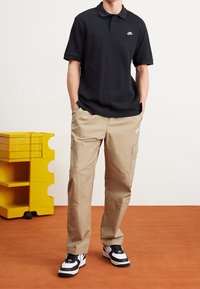 Černé polo tričko s krátkými rukávy a bílým logem Nike, doplněné o béžové cargo kalhoty s kapsami. Oblečení doplňují černo-bílé tenisky.