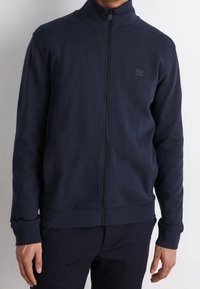Mörkblå zip-up sweatshirt med hög krage, tillverkad av mjukt material; har en diskret logopatch på bröstet och ribbade ärmslut vid handlederna.
