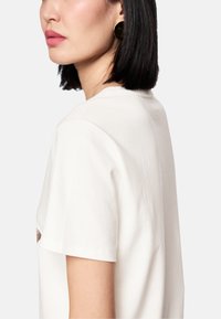 Cremefarbenes Baumwoll-T-Shirt mit kurzen Ärmeln, Rundhalsausschnitt und einer dezenten Struktur, von der Seite gezeigt.