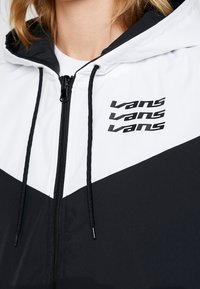 Vit och svart huva jacka med svart dragkedja, snoddar och tre "Vans" logotyper staplade på bröstet, bärs av en person.