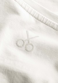 Witte katoenen t-shirt stof met een reliëflogo van scharen, gepositioneerd nabij de halslijn, met een gladde en zachte textuur.