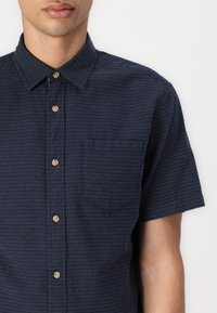 Chemise à manches courtes bleu marine avec un motif géométrique subtil, poche poitrine et boutons beige. Le tissu semble léger et texturé.