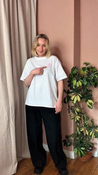 Blonde woman in white t-shirt and black pants points to pink logo, standing next to green potted plant against a pink wall and beige curtain.