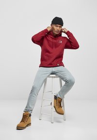 Rød hettegenser med frontlomme og logo, kombinert med lyseblå jeans og brune snørestøvler, sitter på en hvit krakk.