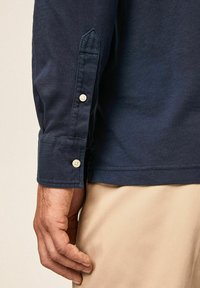 Marineblaues Langarmhemd mit spitzem Manschette, das über einen Knopfverschluss und eine glatte, Baumwolloberfläche verfügt. Hand liegt neben einer beigefarbenen Hose.