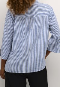 Blue and white striped button-up shirt with three-quarter sleeves, pleated back detail, and a relaxed fit. Cotton fabric texture.