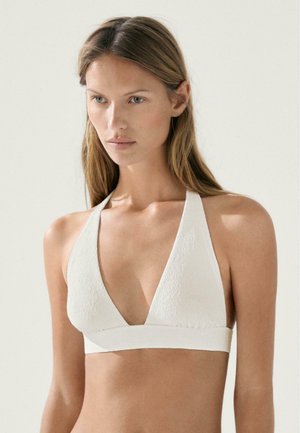 Mladá žena s dlouhými světlými vlasy, která nosí bílé texturované bikiny s halterovým topem na jednobarevném světlém pozadí.