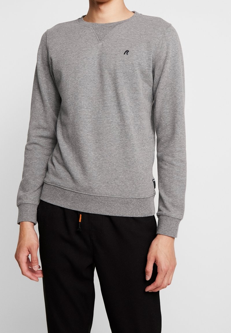 Homme portant un sweat-shirt gris uni à manches longues avec un petit logo noir sur la poitrine et un pantalon noir, sur un fond blanc.