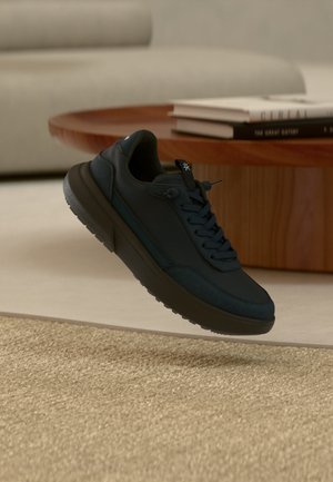 Dunkelblauer Sneaker mit schwarzer Sohle, schwebend über beigem Teppich, einem Holzcouchtisch und gestapelten Büchern in einem zurückhaltenden Wohnzimmer.