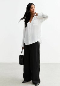 Witte overhemdd, voorzien van een zak, gecombineerd met zwarte wijde broek met witte zijstreep. Een zwarte tas en puntige schoenen maken de look compleet.