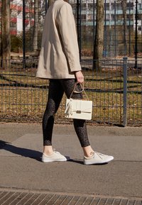 Persona che cammina sul marciapiede indossando una giacca beige, legging a stampa leopardata, sneakers bianche e portando una borsa bianca con catena dorata.