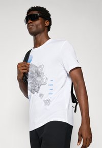 Hvid bomulds t-shirt med et sort konturdesign og blå tekst. Korte ærmer og rund halsudskæring. Sort rygsækrem er synlig.