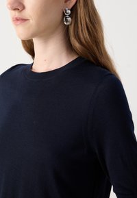 Femme aux longs cheveux châtain clair portant un pull bleu marine et de grandes boucles d'oreilles doubles boules argentées réfléchissantes.
