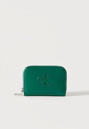 Portefeuille en cuir vert avec fermeture éclair, logo "cK" embossé sur le devant et tirette en métal argenté, posé sur un fond clair uni.