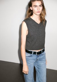 Donkergrijze mouwloze gebreide trui met een V-hals, gecombineerd met lichtblauwe high-waisted jeans en een zwarte riem met een zilveren gesp.