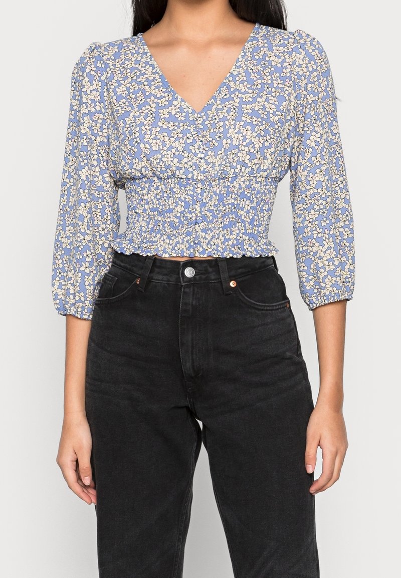 Femme portant un chemisier smocké bleu à fleurs avec un col en V, des manches trois-quarts et un jean noir taille haute, debout devant un fond uni.