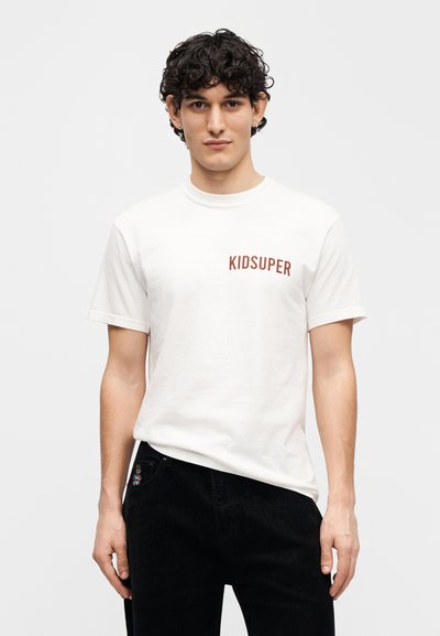 Λευκό βαμβακερό μπλουζάκι με τη λέξη "KIDSUPER" κόκκινη στο αριστερό μέρος του στήθους. Κοντά μανίκια, στρογγυλή λαιμόκοψη, απλός σχεδιασμός.