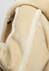 Beige fleecejakke med en blød tekstur, cremefarvet foring og en rund krave med en træknapdetalje.